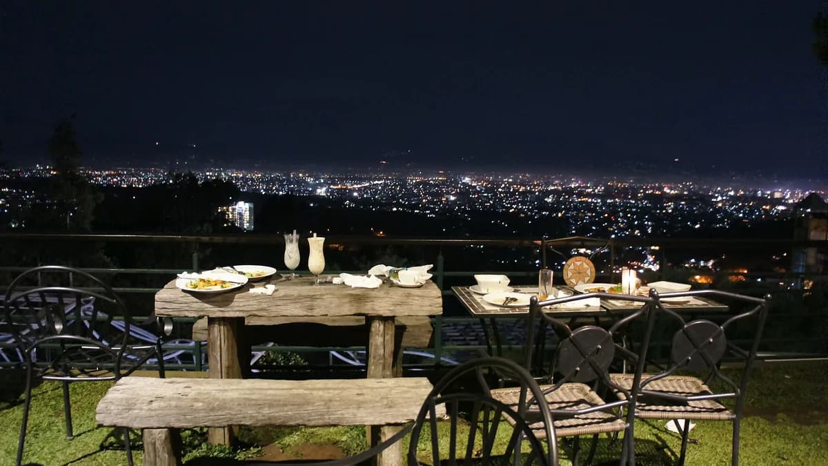 Lampu kota Bandung dilihat dari teras restoran di perbukitan Dago pada malam hari, dengan meja makan kayu di latar depan