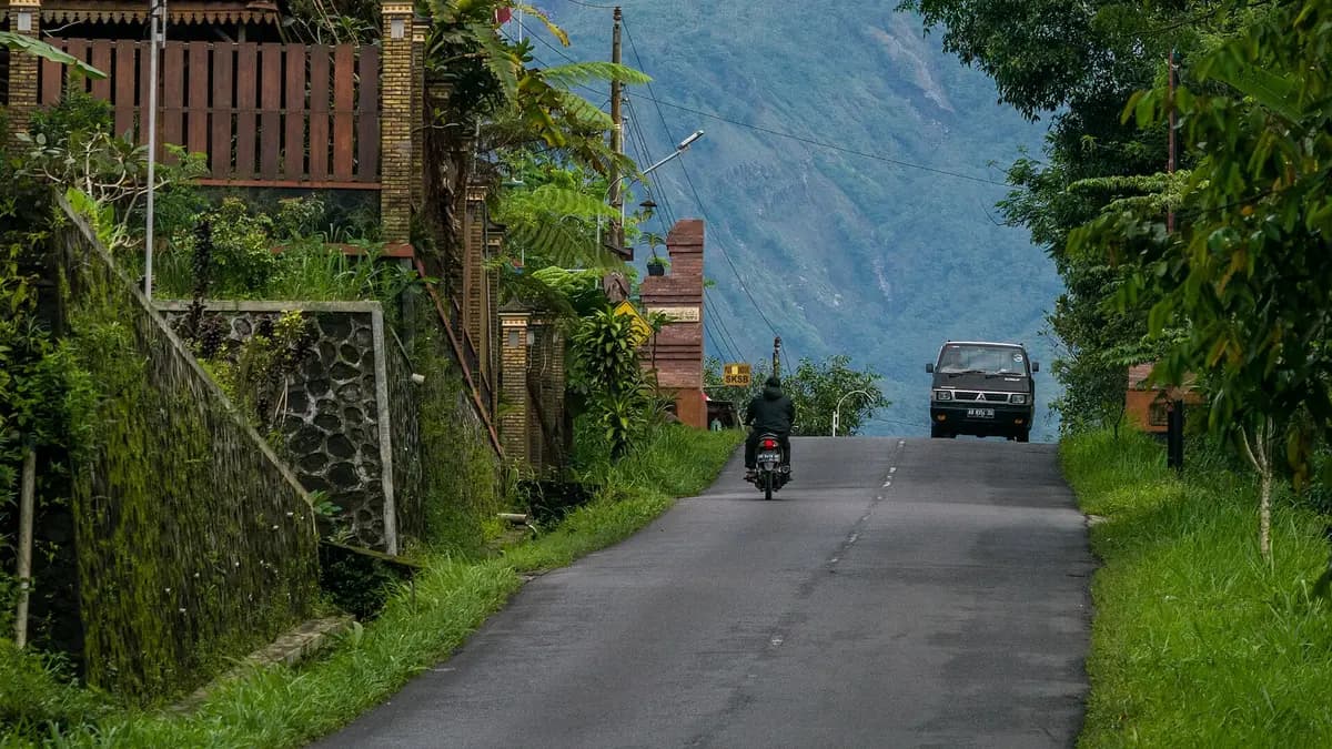 Jalan desa menanjak di Kalurahan Kepuharjo, Cangkringan, dengan pengendara motor dan mobil bak di depan lereng Merapi