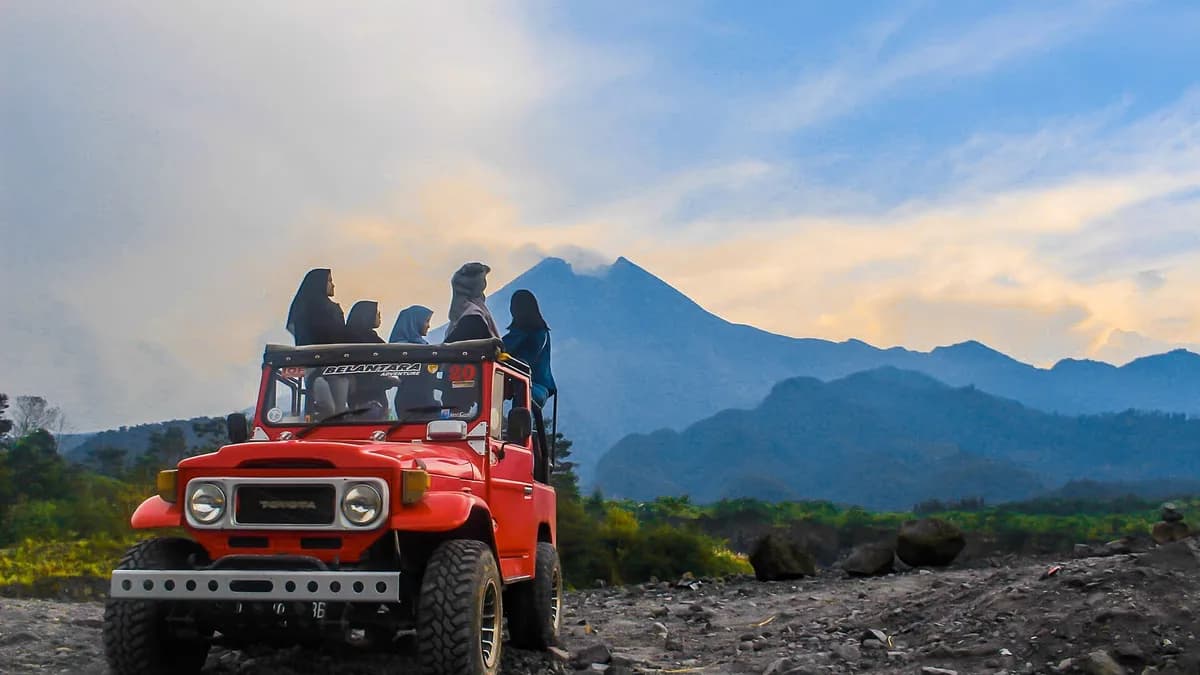 Jeep merah terbuka membawa rombongan wisatawan di jalur berbatu Lava Tour Merapi dengan punggungan gunung di latar