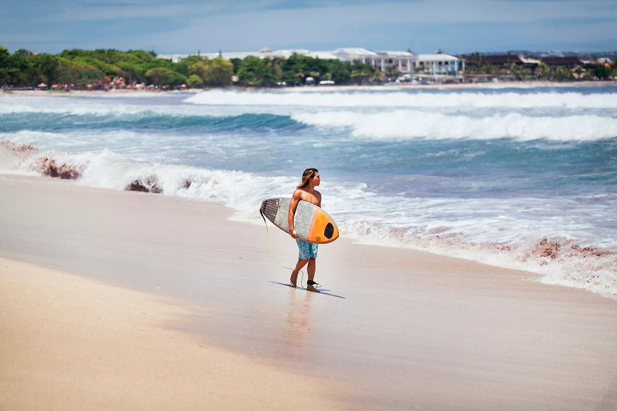 Peselancar menjinjing papan berjalan di tepi air Pantai Kuta Bali dengan ombak bergulung di belakangnya