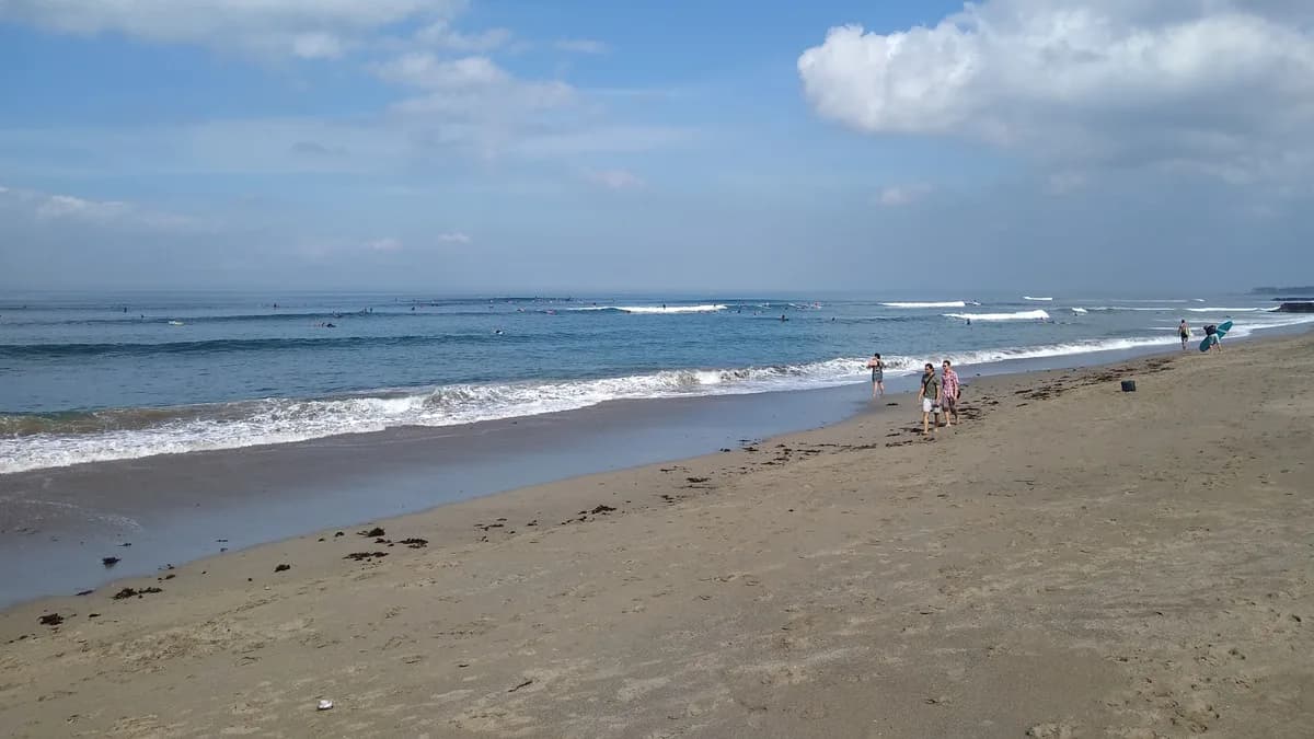Ombak bergulung di Pantai Canggu dengan pasir gelap dan beberapa peselancar di kejauhan