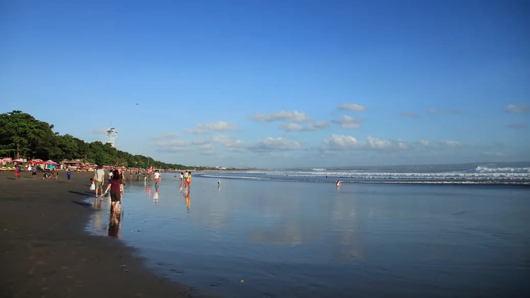 Garis Pantai Kuta yang panjang dengan pengunjung berjalan di pasir basah dan ombak kecil di kejauhan