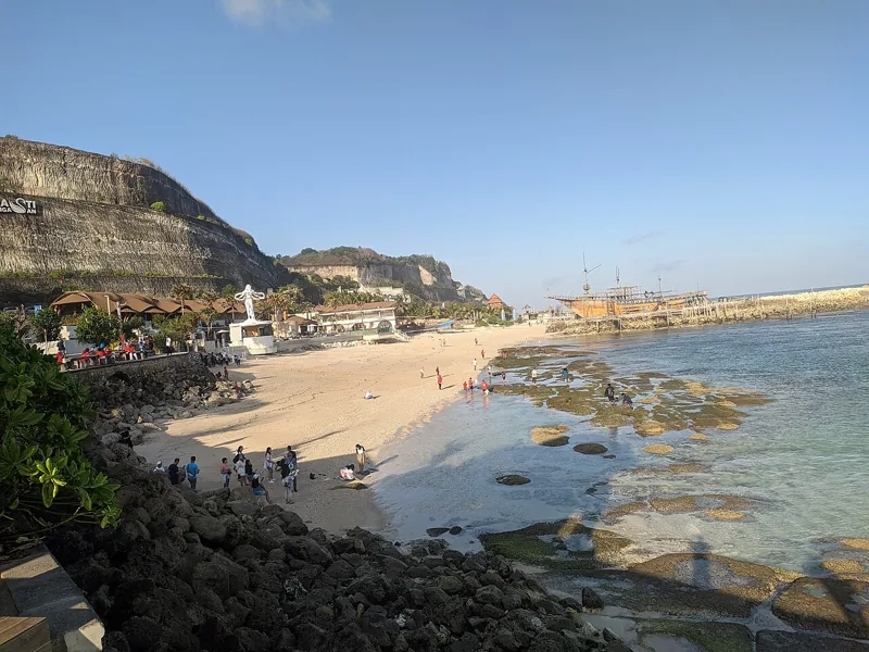 Pantai Melasti dilihat dari samping: pasir putih, karang yang tersingkap saat surut, tebing kapur bertuliskan Melasti Ungasan, dan replika kapal kayu di ujung pantai