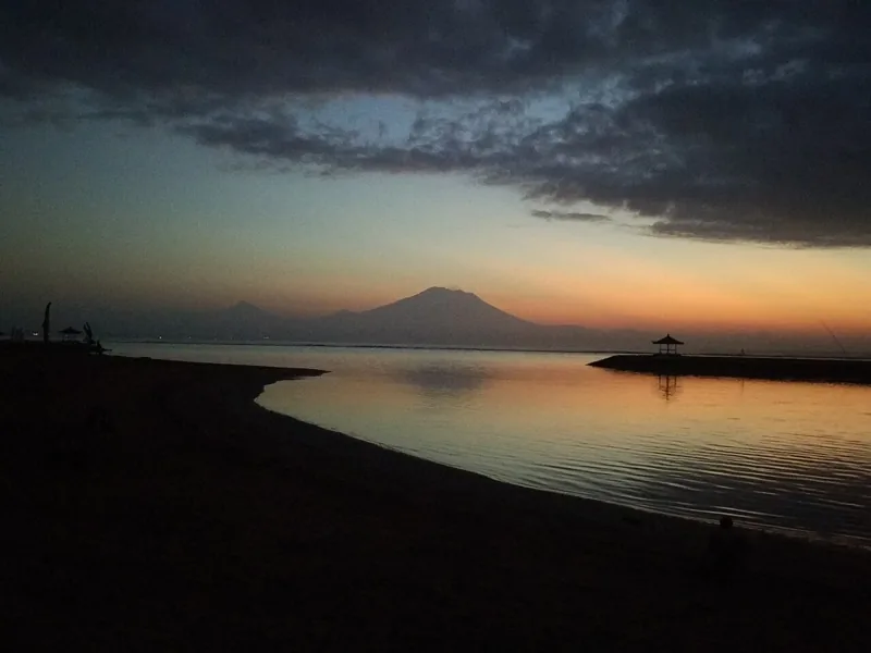 Langit fajar jingga di tepi Pantai Sanur, dengan siluet gazebo di ujung pemecah ombak dan gunung di cakrawala