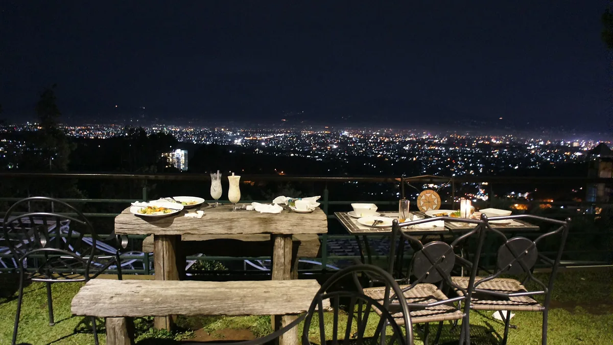 Lampu kota Bandung dilihat dari teras restoran di perbukitan Dago pada malam hari, dengan meja makan kayu di latar depan