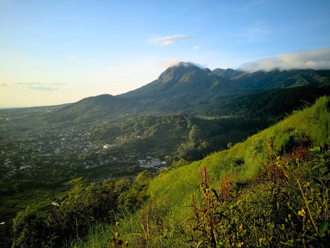 Lereng berumput di Kota Batu pada pagi hari dengan Gunung Panderman berselimut awan dan permukiman kota di bawahnya