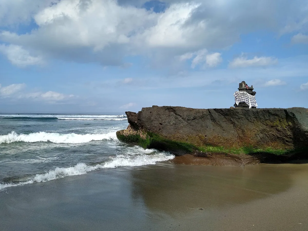 Karang dengan pelinggih kecil di bibir Pantai Canggu dengan ombak pecah putih di belakangnya