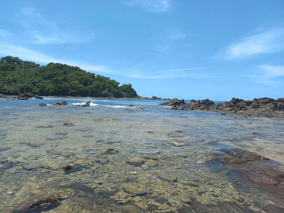 Air laut dangkal yang jernih di atas hamparan karang dengan tanjung berhutan di kejauhan