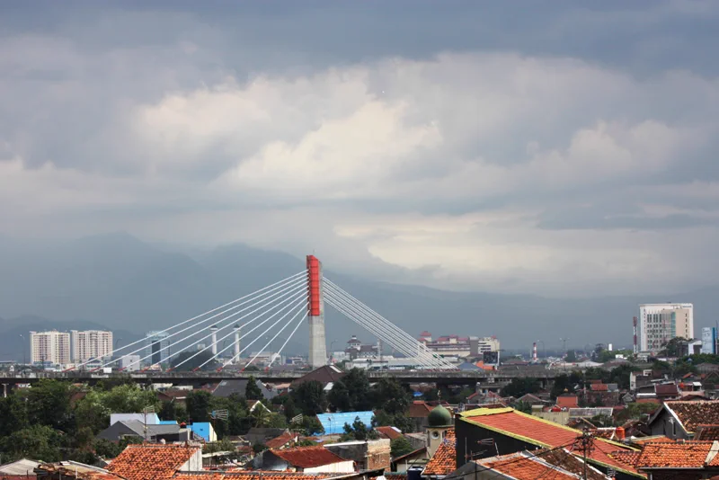 Menara merah putih Jembatan Layang Pasupati menonjol di atas atap genteng permukiman Bandung dengan punggungan gunung di kejauhan