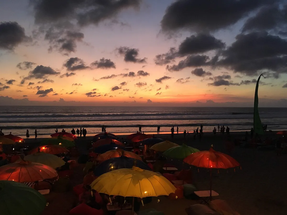 Payung-payung warna-warni kafe pantai Seminyak dengan langit jingga saat matahari terbenam