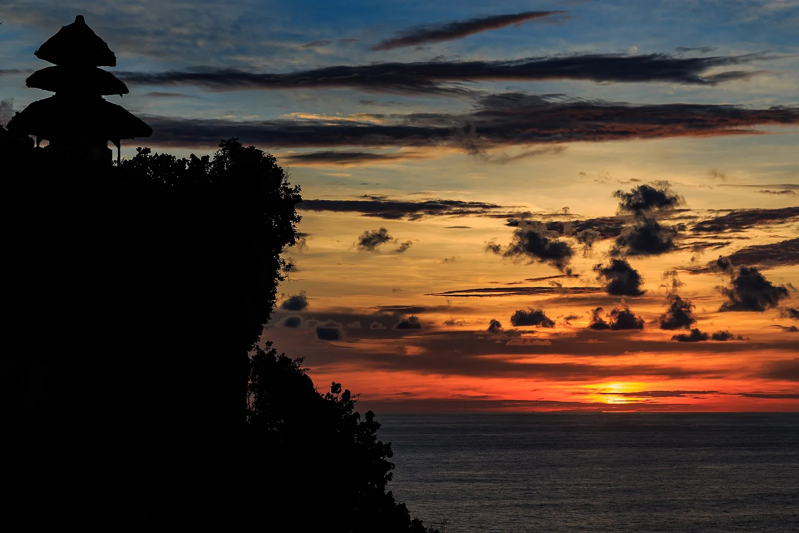 Siluet meru Pura Luhur Uluwatu di ujung tebing dengan matahari terbenam di atas Samudra Hindia