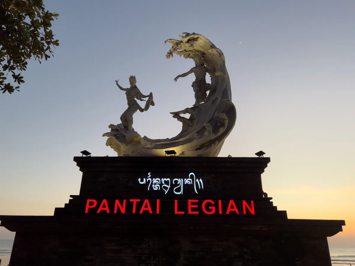Gapura bertuliskan PANTAI LEGIAN dengan patung di atasnya, difoto saat langit senja