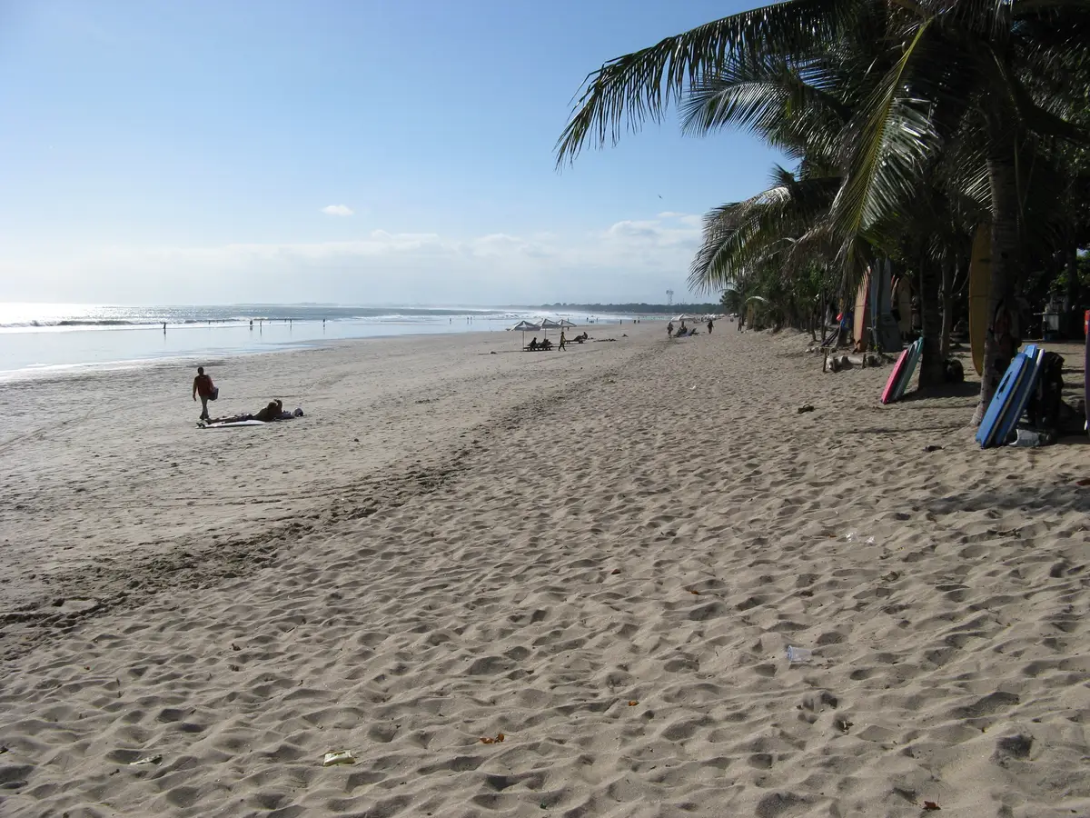 Hamparan pasir Pantai Legian yang lebar dengan pohon kelapa di tepi dan pengunjung berjalan di dekat ombak