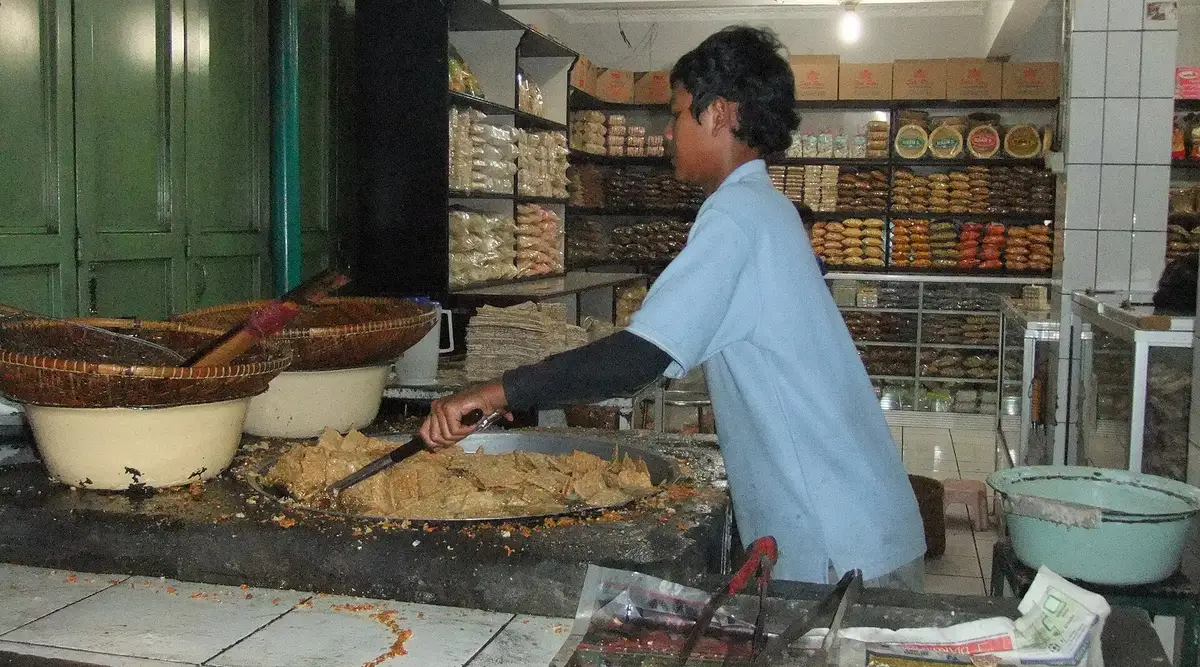 Penjual menggoreng keripik tempe di sebuah toko jajanan di Lembang dengan rak kemasan oleh-oleh di belakangnya