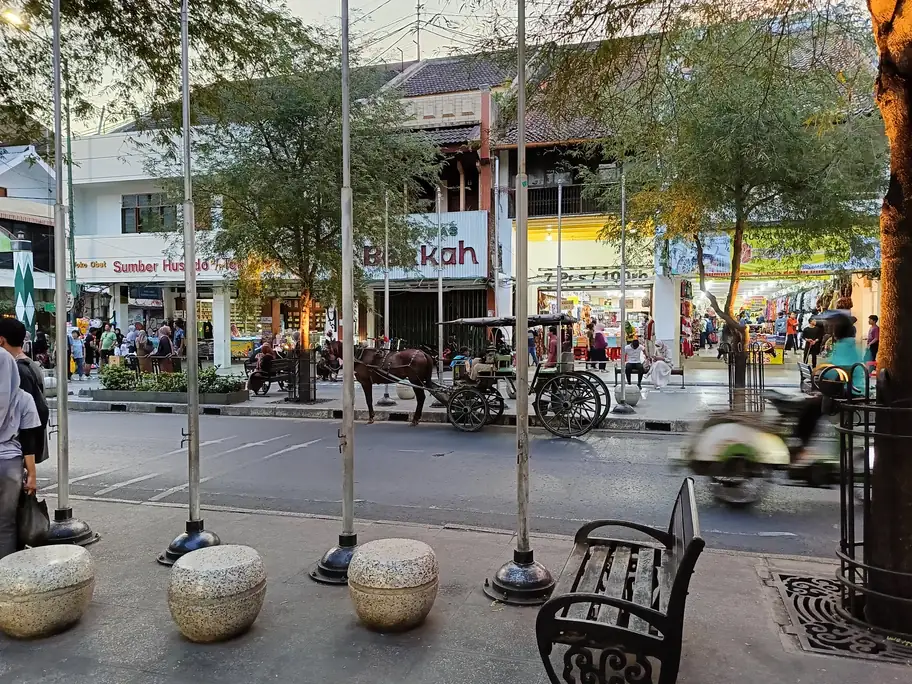 Andong dan becak menunggu penumpang di tepi Jalan Malioboro saat senja, dengan pejalan kaki dan kios oleh-oleh di sekitarnya
