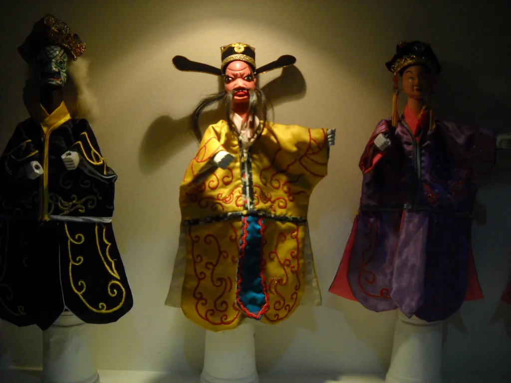 Tiga boneka wayang potehi berkostum sutra kuning, hitam, dan ungu dipajang berjajar di dalam vitrin Museum Wayang