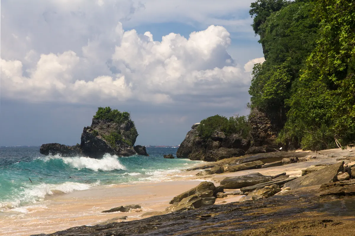 Gugusan karang dan ombak putih di sisi Pantai Padang Padang dengan tebing berhutan di latar belakang