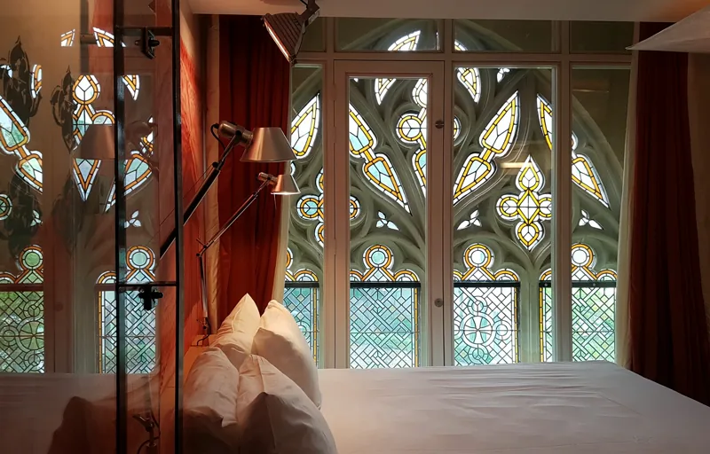 Kamar hotel dengan ranjang putih menghadap deretan jendela kaca patri bekas bangunan gereja