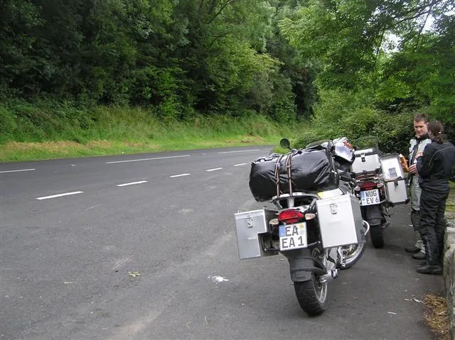 Dua motor touring bermuatan tas dan boks parkir di bahu jalan sementara dua pengendaranya beristirahat di tepi hutan