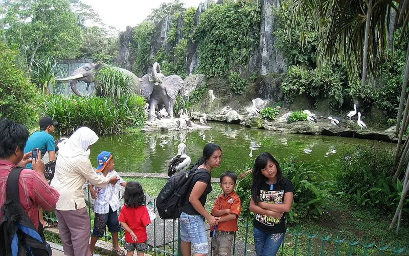Orang tua dan anak-anak berdiri di tepi kolam pelikan Taman Margasatwa Ragunan dengan patung gajah di latar belakang