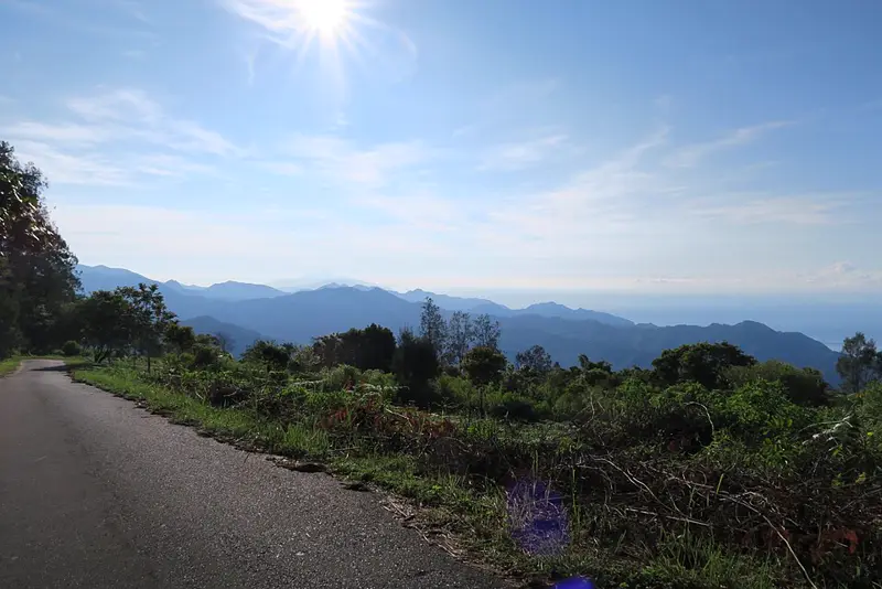 Jalan beraspal menurun dari kawasan Gunung Kelimutu dengan semak hijau dan deretan perbukitan biru di kejauhan