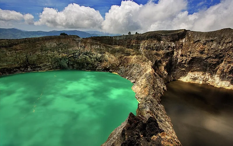 Dua danau kawah Kelimutu bersebelahan, satu hijau toska dan satu cokelat gelap, dipisahkan dinding kawah