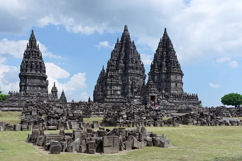 Menara-menara runcing Candi Prambanan menjulang di bawah langit berawan dengan tumpukan batu reruntuhan di halaman