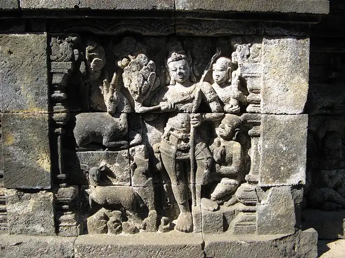 Panel relief batu Candi Borobudur berisi tokoh pemburu membawa busur bersama pengiring dan hewan di tengah adegan hutan