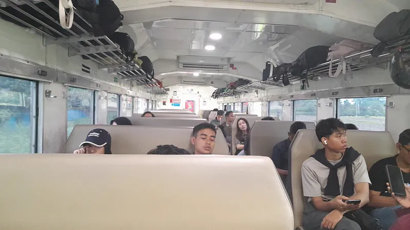Interior kereta kelas ekonomi dengan kursi berhadapan, rak bagasi penuh ransel, dan penumpang muda