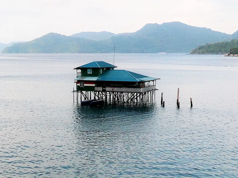 Bangunan kayu beratap hijau berdiri di atas tiang-tiang di tengah perairan tenang dengan latar pulau berbukit