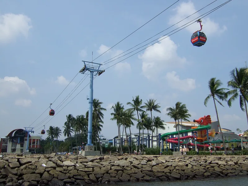 Kabin gondola merah melintas pada kabel yang ditopang tiang biru di tepi pantai Ancol dengan deretan pohon kelapa