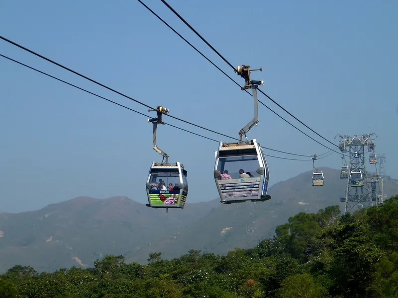 Dua kabin gondola berisi penumpang bergantung pada kabel di atas hutan dengan latar perbukitan, Ngong Ping 360 Hong Kong