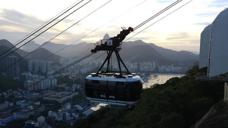 Kabin kereta gantung Pao de Acucar bergantung pada kabel dengan latar kota Rio de Janeiro, teluk, dan pegunungan saat senja