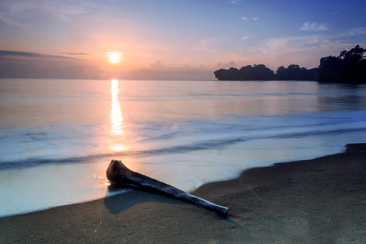 Matahari terbit di Pantai Batu Karas dengan sebatang kayu terdampar di pasir dan tanjung berhutan di kejauhan