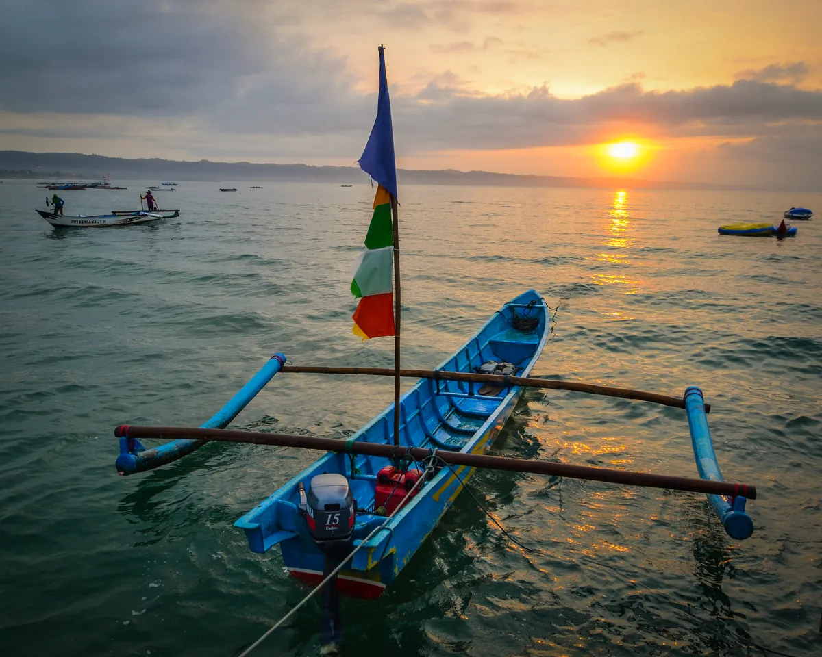 Perahu jukung bercadik dengan bendera warna-warni mengapung di Pantai Timur Pangandaran saat matahari terbit
