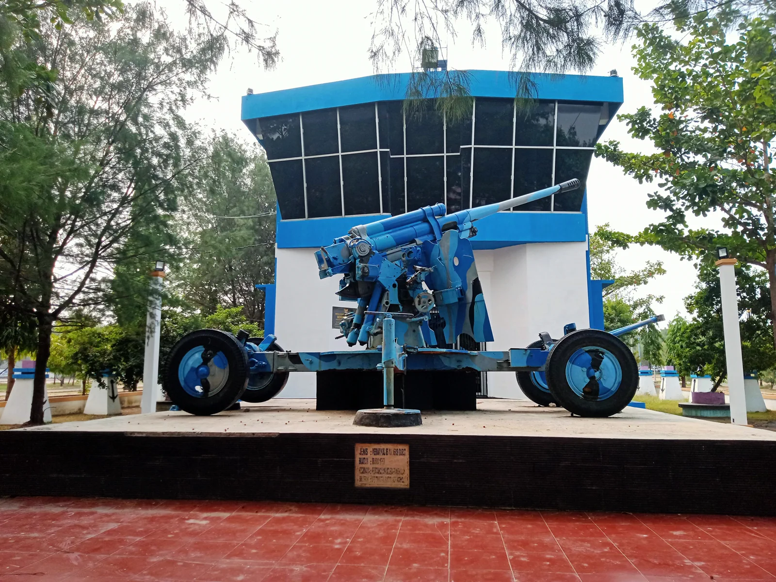 Monumen Bahari berbentuk anjungan kapal berwarna biru putih dengan meriam penangkis serangan udara di depannya, dikelilingi pohon cemara laut