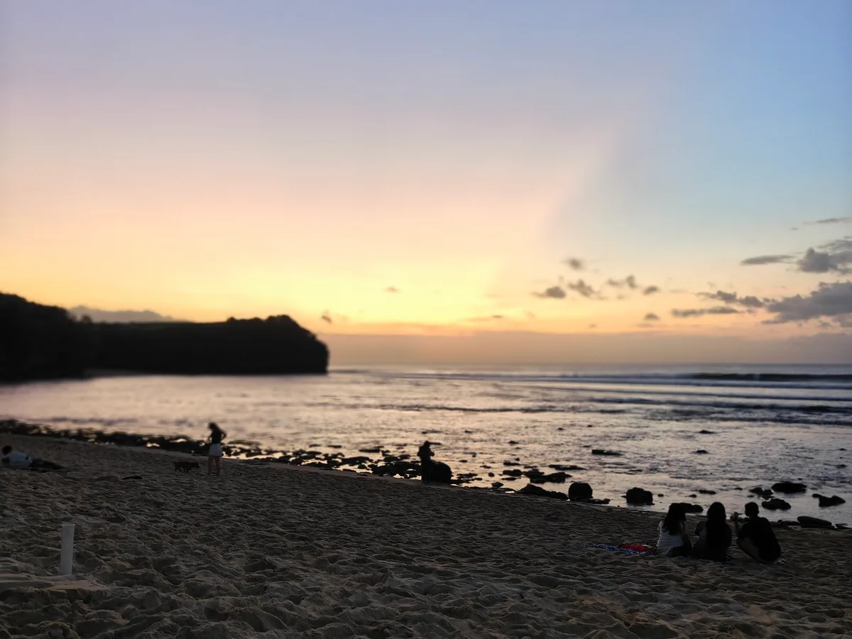 Matahari terbenam di Pantai Balangan, Kuta Selatan, dengan siluet tebing dan pengunjung duduk di pasir