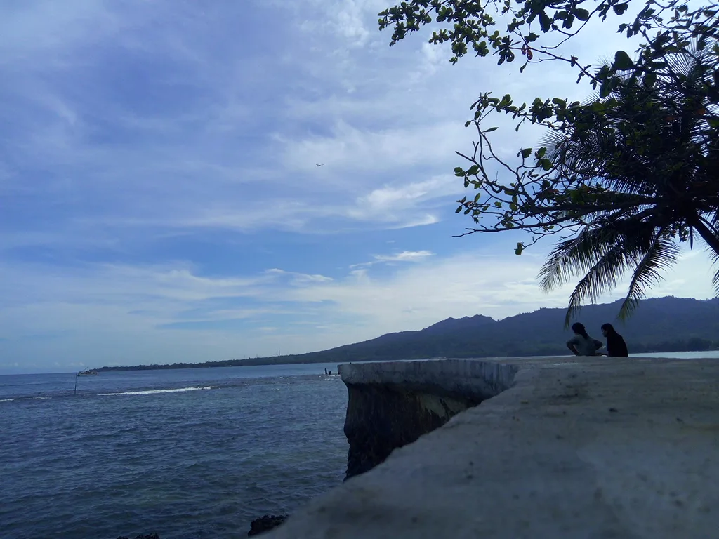 Dua orang duduk di tanggul beton tepi Pantai Carita di bawah pohon kelapa miring dengan punggungan bukit di kejauhan