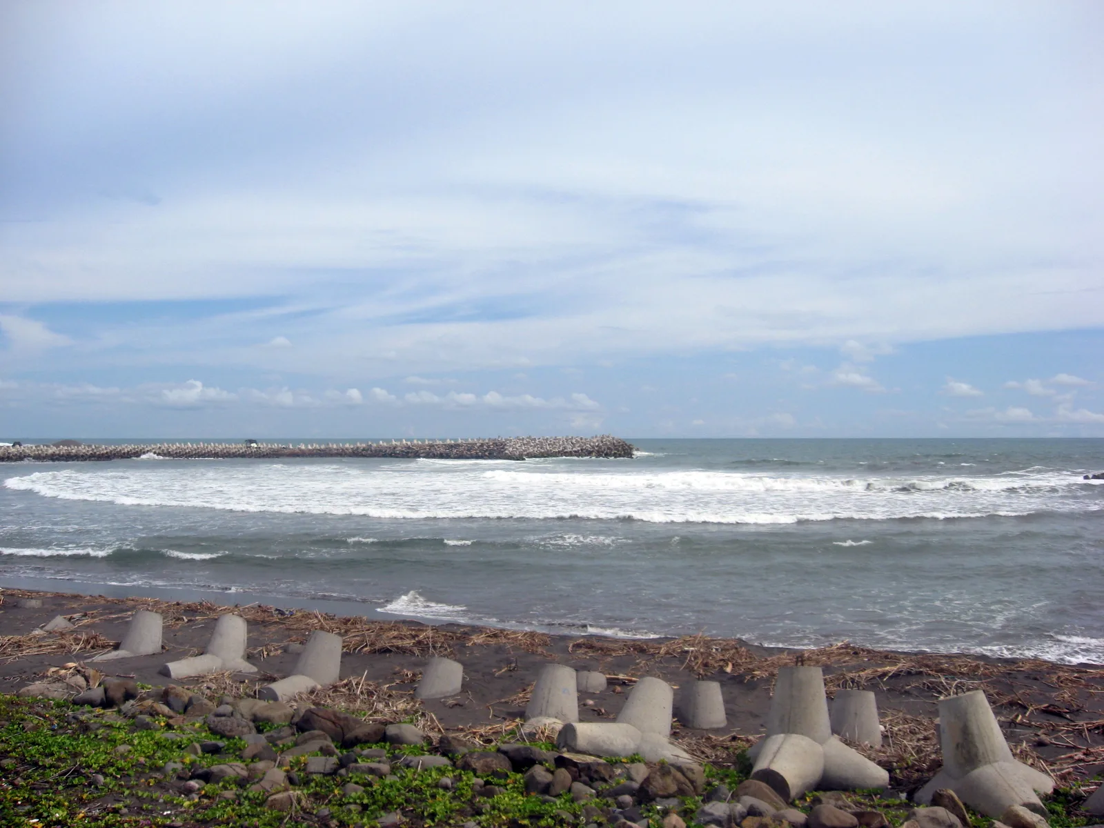 Barisan tetrapod beton di pasir gelap Pantai Glagah dengan pemecah ombak batu memanjang ke laut