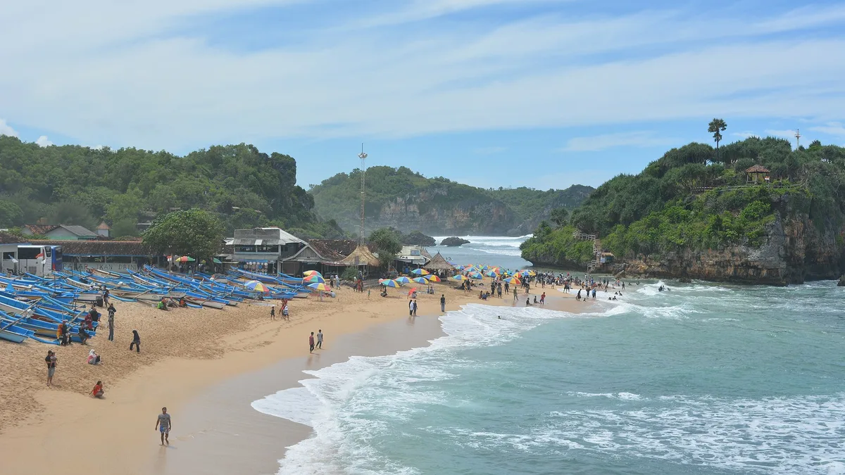 Pantai Drini dilihat dari atas, dengan deretan perahu nelayan biru di pasir, payung sewa, dan bukit karang di kedua sisi teluk