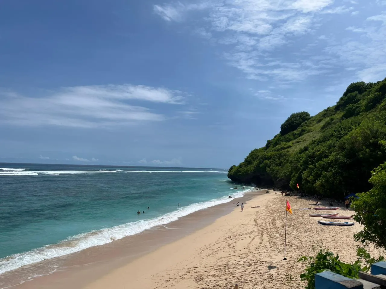 Hamparan pasir putih Pantai Gunung Payung dengan air laut kehijauan, bendera penjaga pantai, dan papan selancar di sisi tebing