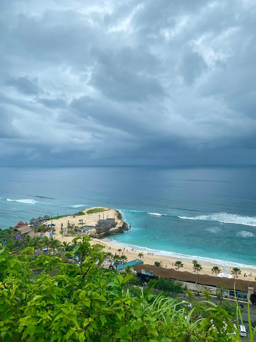 Pemandangan Pantai Melasti Ungasan dari atas tebing, memperlihatkan tanjung berpasir yang menjorok ke laut, deretan kelab pantai, dan air biru kehijauan