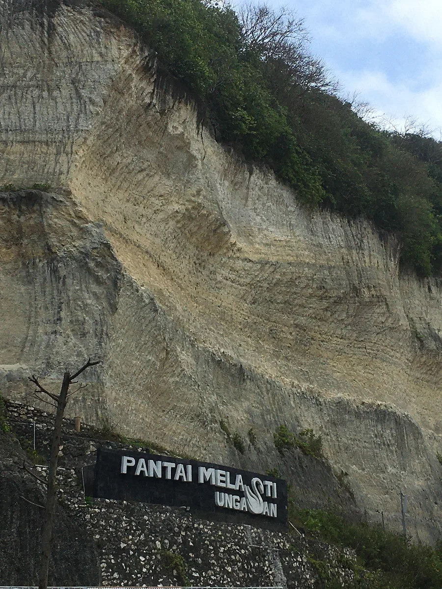 Papan nama besar bertuliskan Pantai Melasti Ungasan menempel di kaki tebing kapur berlapis di jalan menuju pantai