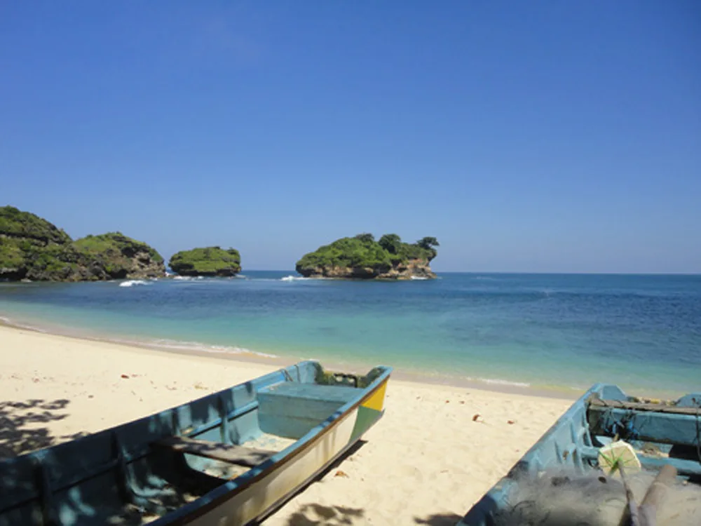 Perahu nelayan tersandar di pasir putih Pantai Watu Karung dengan tiga karang berpuncak hijau di lepas pantai
