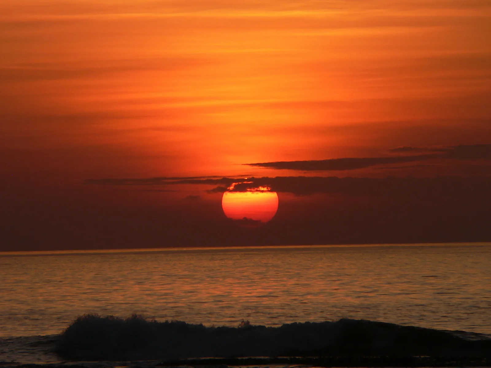 Matahari terbit merah di atas laut dilihat dari Pantai Saba dengan satu ombak pecah di garis karang