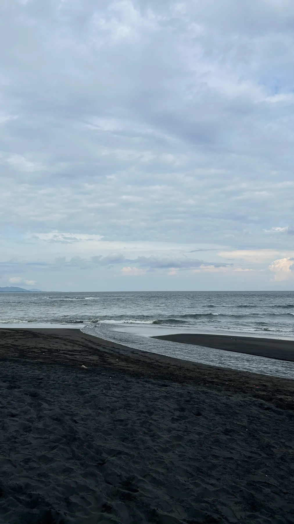 Hamparan pasir hitam Pantai Saba yang basah dengan ombak kecil dan garis bukit samar di kejauhan