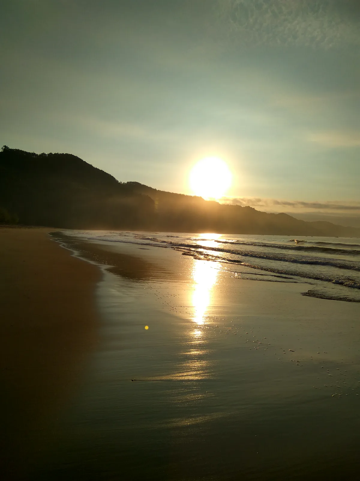 Matahari terbit di atas laut di Pantai Sine dengan pasir basah memantulkan cahaya dan punggungan bukit gelap di sisi kiri