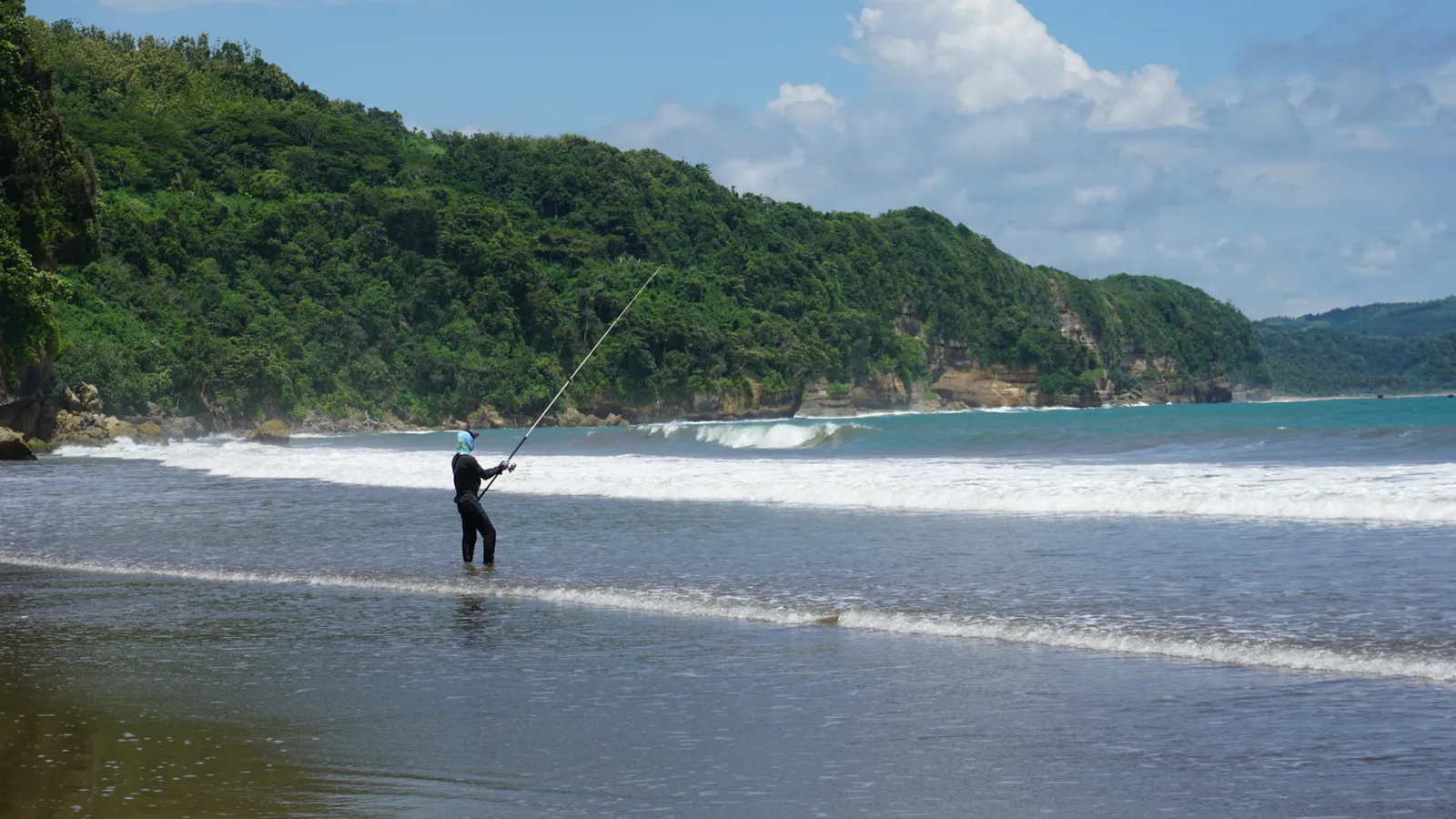 Seorang pemancing berdiri di air dangkal Pantai Sine sambil melempar joran, dengan tebing berhutan hijau di belakangnya
