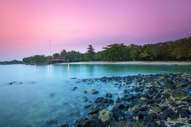Pantai Tanjung Lesung saat fajar dengan langit merah muda, batu hitam di tepi air, dan pepohonan rimbun di sepanjang garis pantai