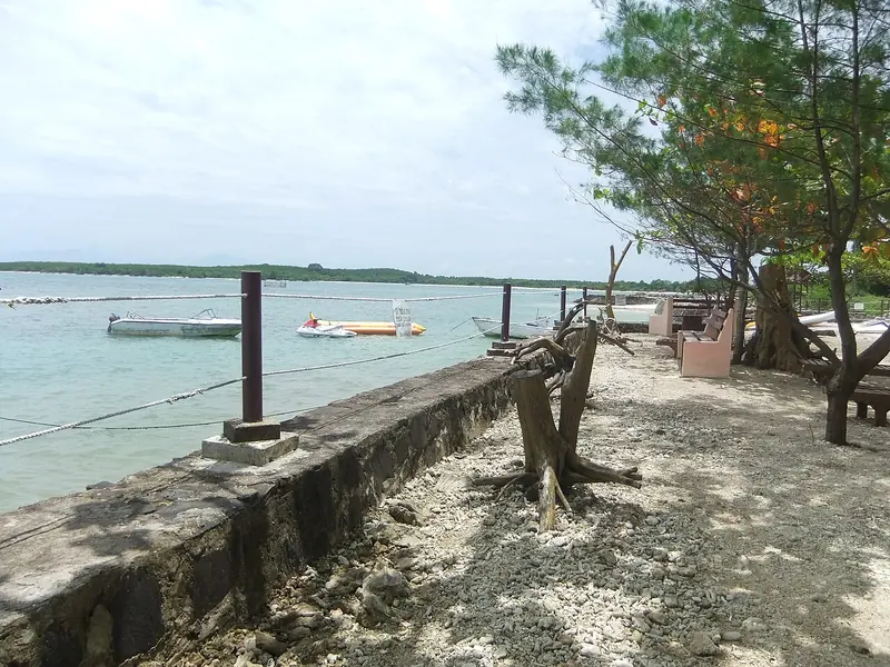 Jalan setapak berbatu di tepi pantai Tanjung Lesung dengan bangku taman, pohon cemara laut, perahu motor, dan banana boat tertambat di air dangkal